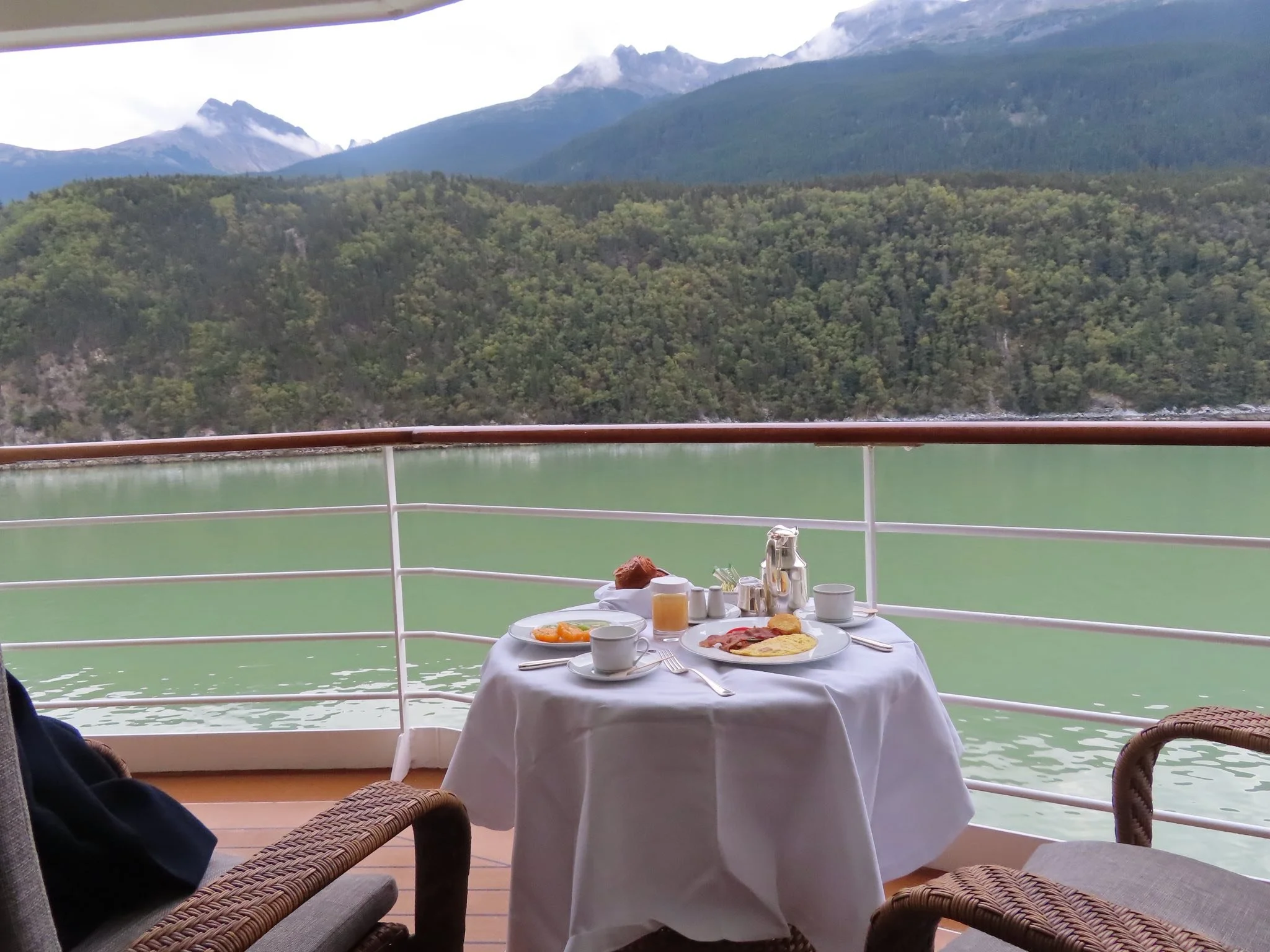 Breakfast table set on a balcony with scenic mountain view in the background, including plates of eggs, bacon, croissants, and beverages.