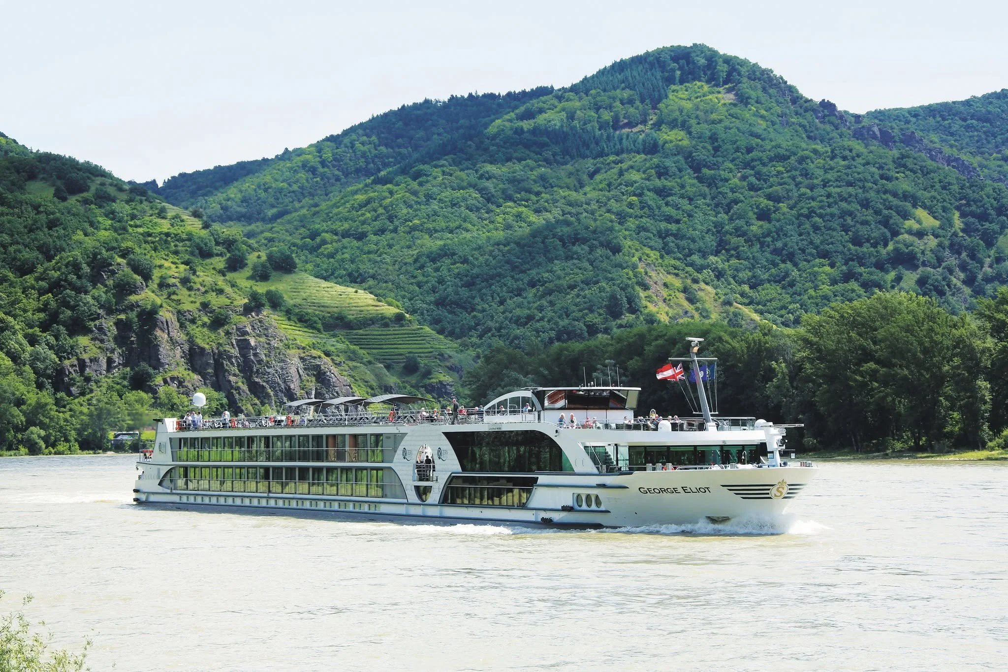 A large white river cruise ship named George Eliot sailing on a river, with green mountains and lush vegetation in the background.