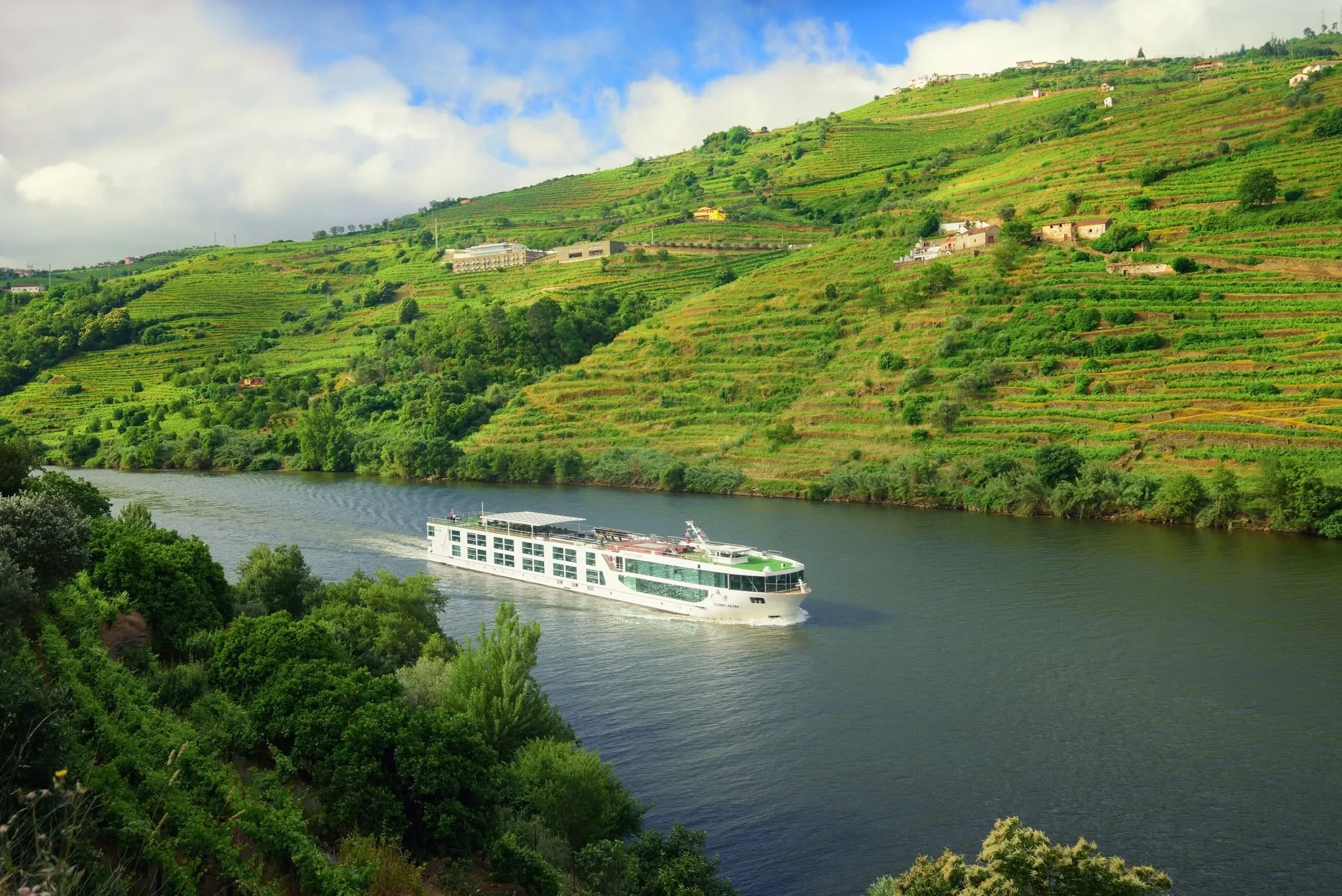 A river running through a lush green valley with hills covered in terraced vineyards and small houses. A white cruise ship is sailing on the river under a partly cloudy sky.