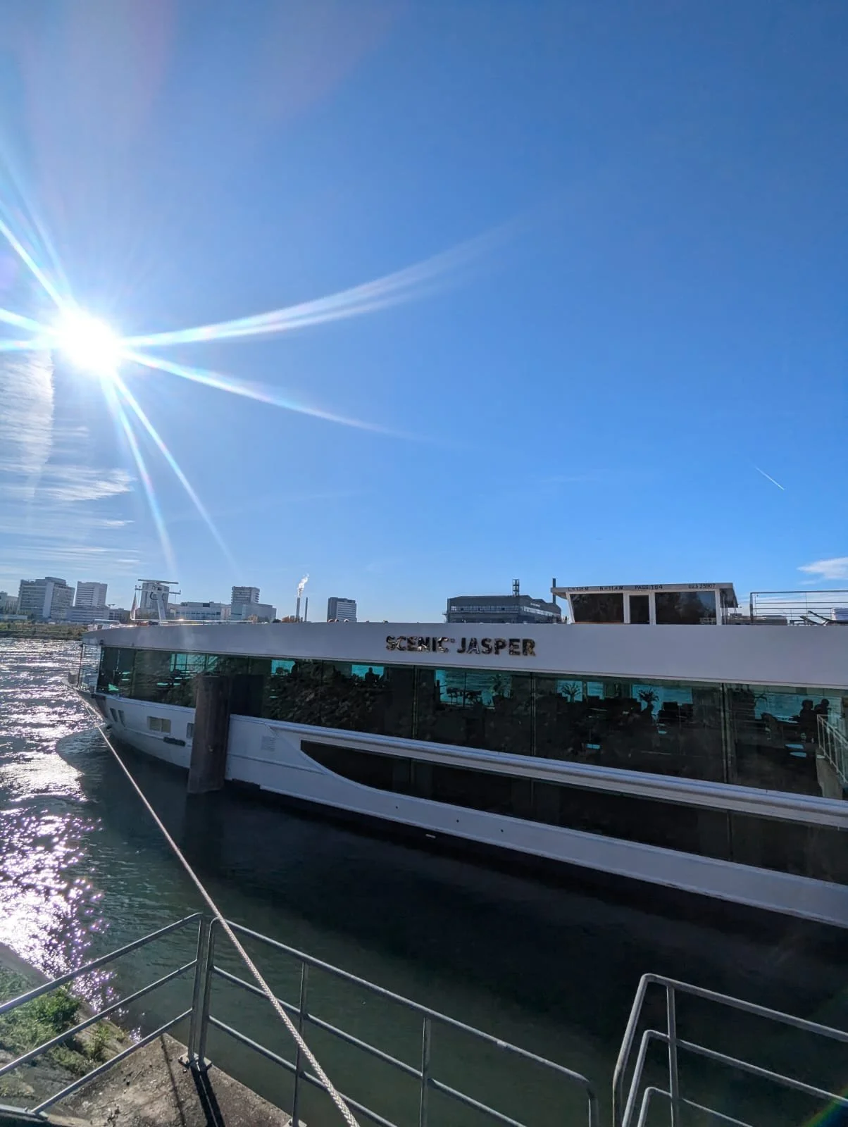 View of a modern white boat named 'Scenic Jasper' docked on a river under a bright sunny sky with some city buildings in the background.