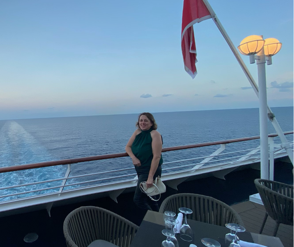 Woman smiling on a cruise ship deck near the railing with the ocean in the background, a red and white flag, and a lit lamp post.