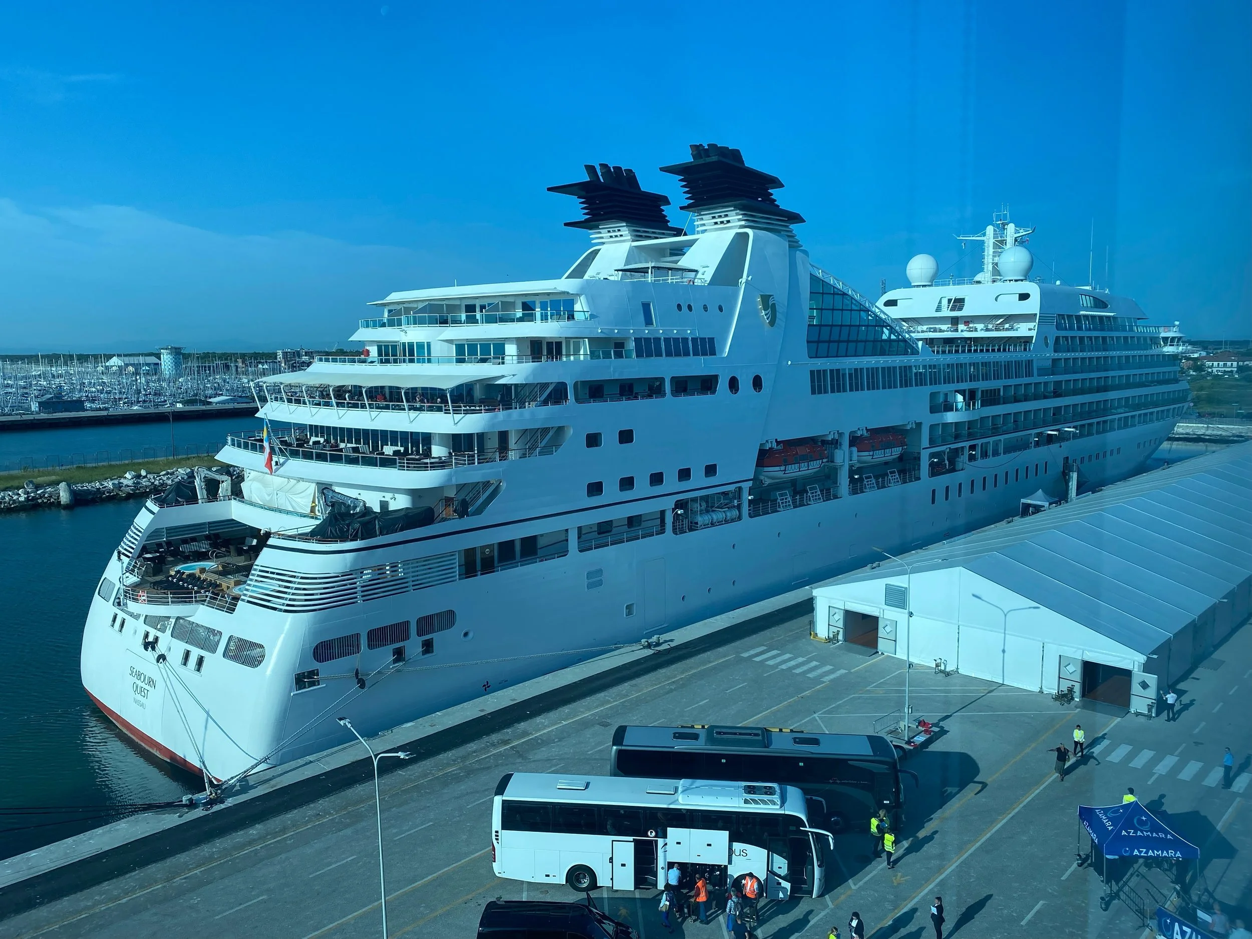 A large white cruise ship docked at a port under a clear blue sky, with several people and vehicles on the dock.
