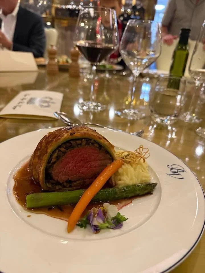 A plated Beef Wellington with mashed potatoes, a green bean, and a carrot, garnished with edible flowers, on a white plate in a restaurant setting with wine glasses and people in the background.