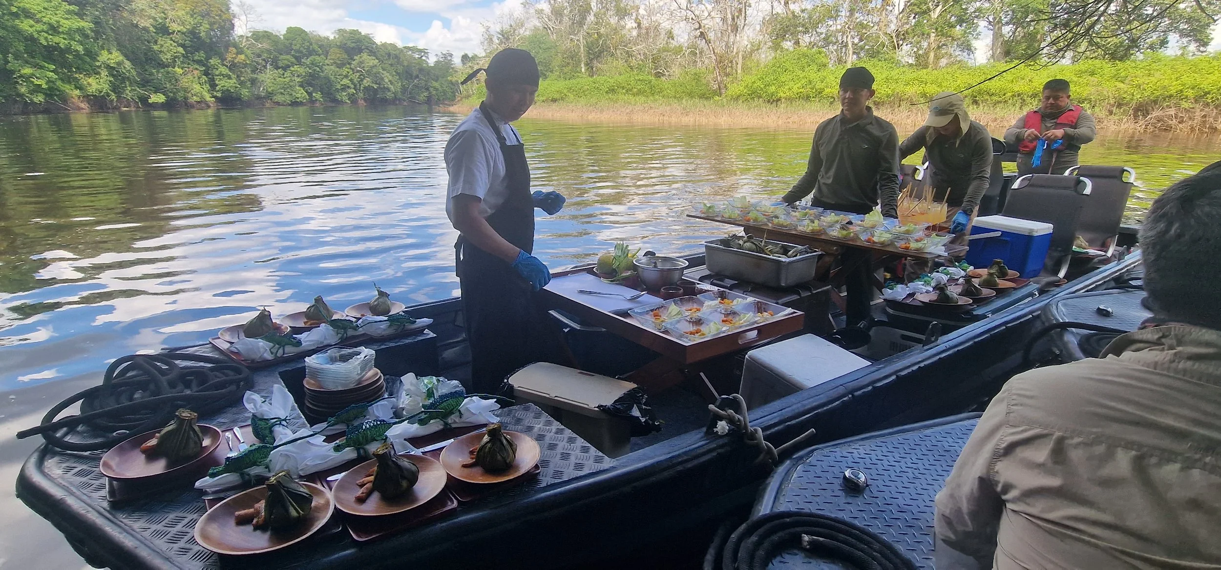 A group of people on a boat by a river, preparing and serving food that includes dishes wrapped in leaves, with a lush green forest in the background.