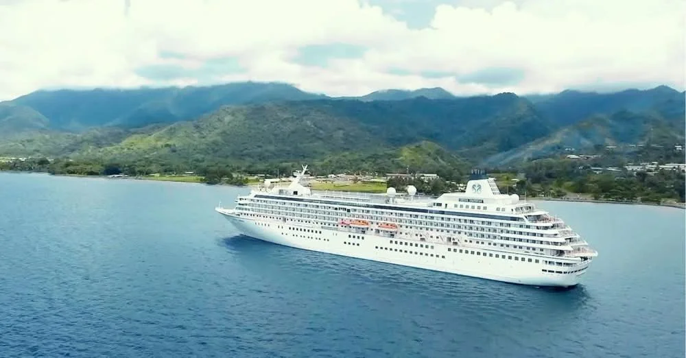 A white cruise ship sailing near a lush green mountainous coastline under a partly cloudy sky.