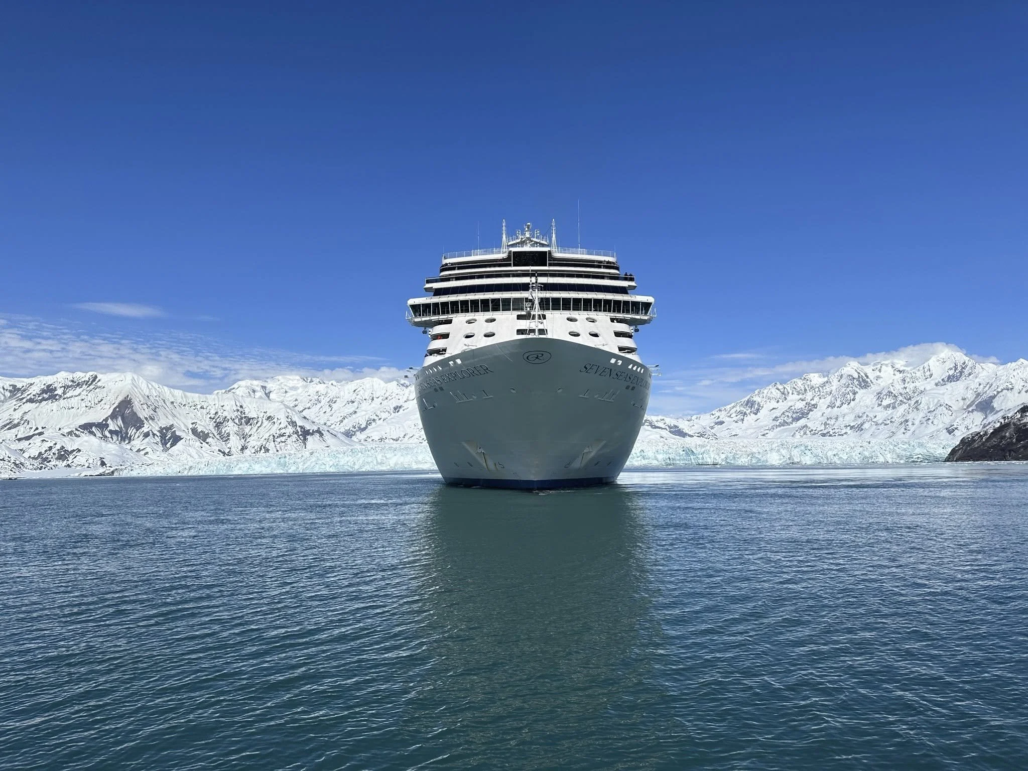 A large white cruise ship moving through icy waters with snow-capped mountains in the background under a clear blue sky.