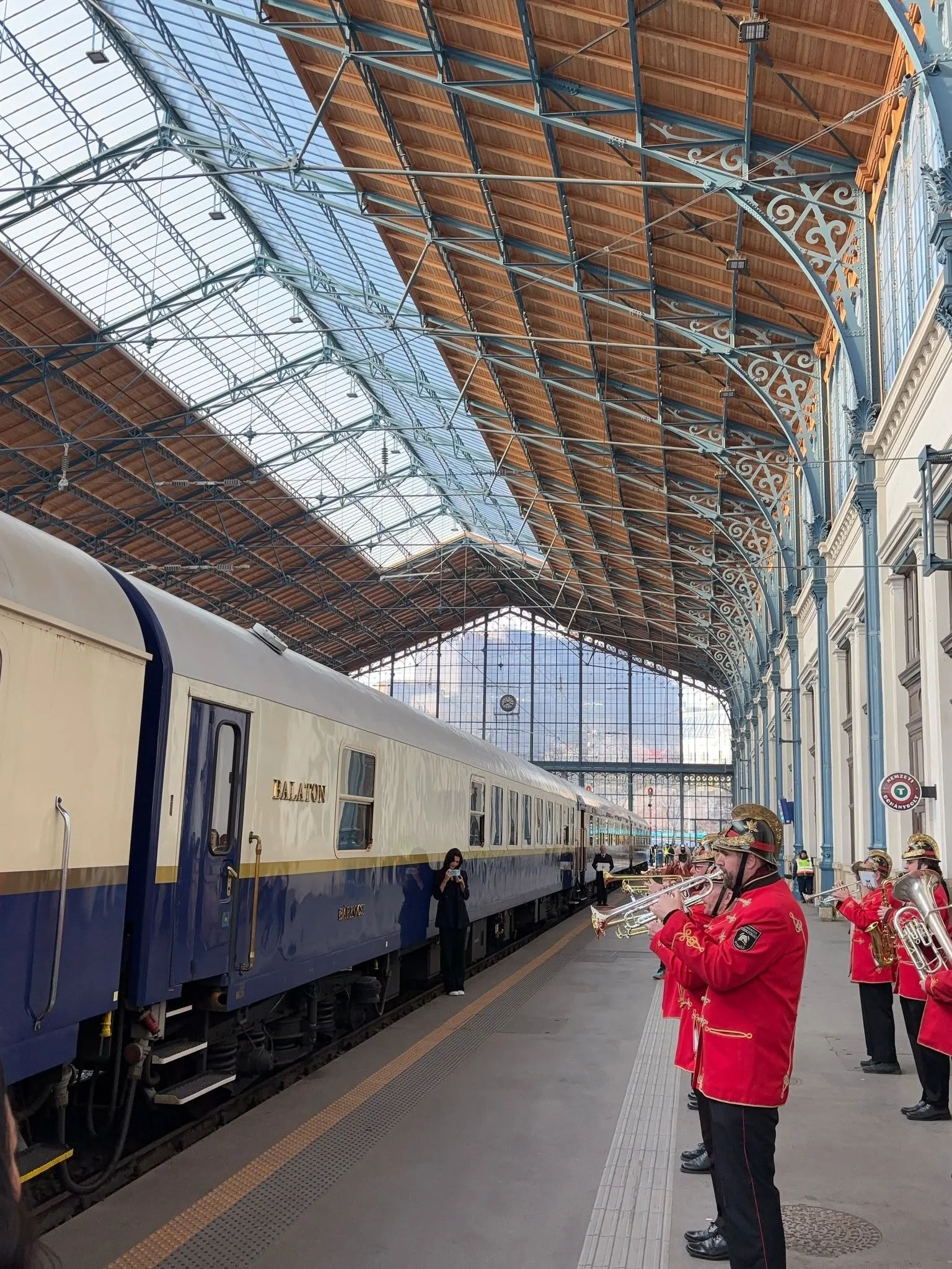 People in ceremonial uniforms playing musical instruments on a train platform with a large glass and metal roof overhead.