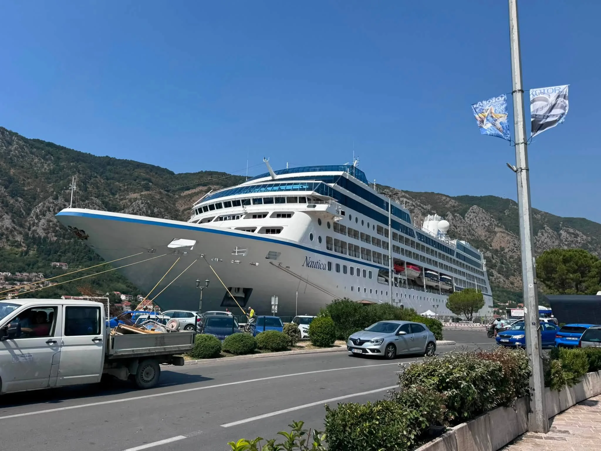 Large cruise ship docked at a port with cars parked in front, a mountain range in the background, and clear blue sky.