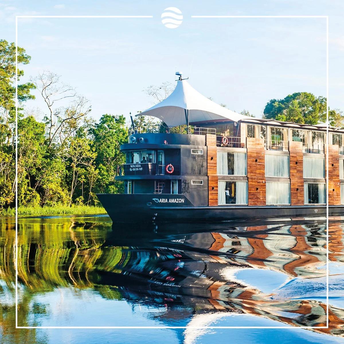 A luxury river ship on calm water surrounded by lush green trees, with reflections visible on the water's surface.