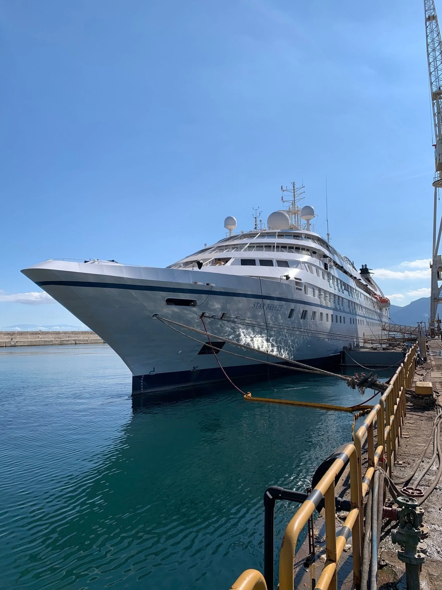 A large white luxury yacht named 'Star Breeze' is docked at a marina with calm water, under a bright blue sky with a few clouds. The boat is tied to the dock with yellow ropes, and there is a safety railing along the pier.