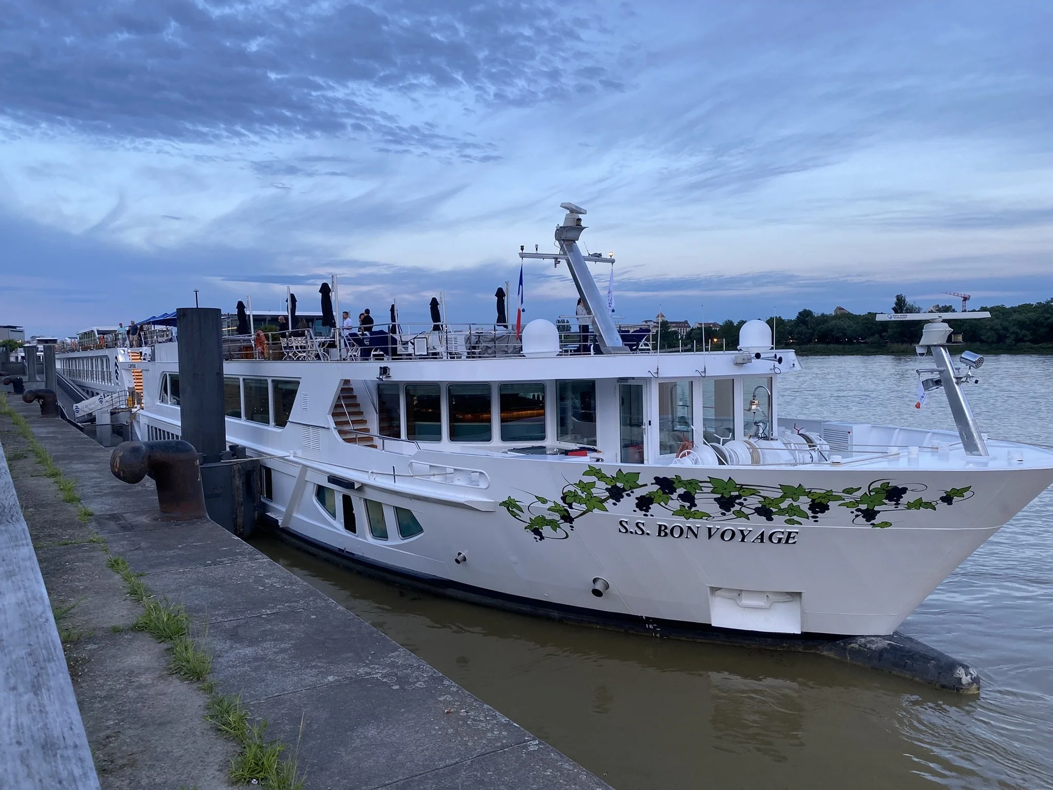 A white boat named S.S. Bon Voyage docked at a pier on a river, with a cityscape and cranes in the background, under a partly cloudy sky.