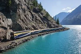 A train traveling along a narrow railway track beside a large body of water in a mountainous area.