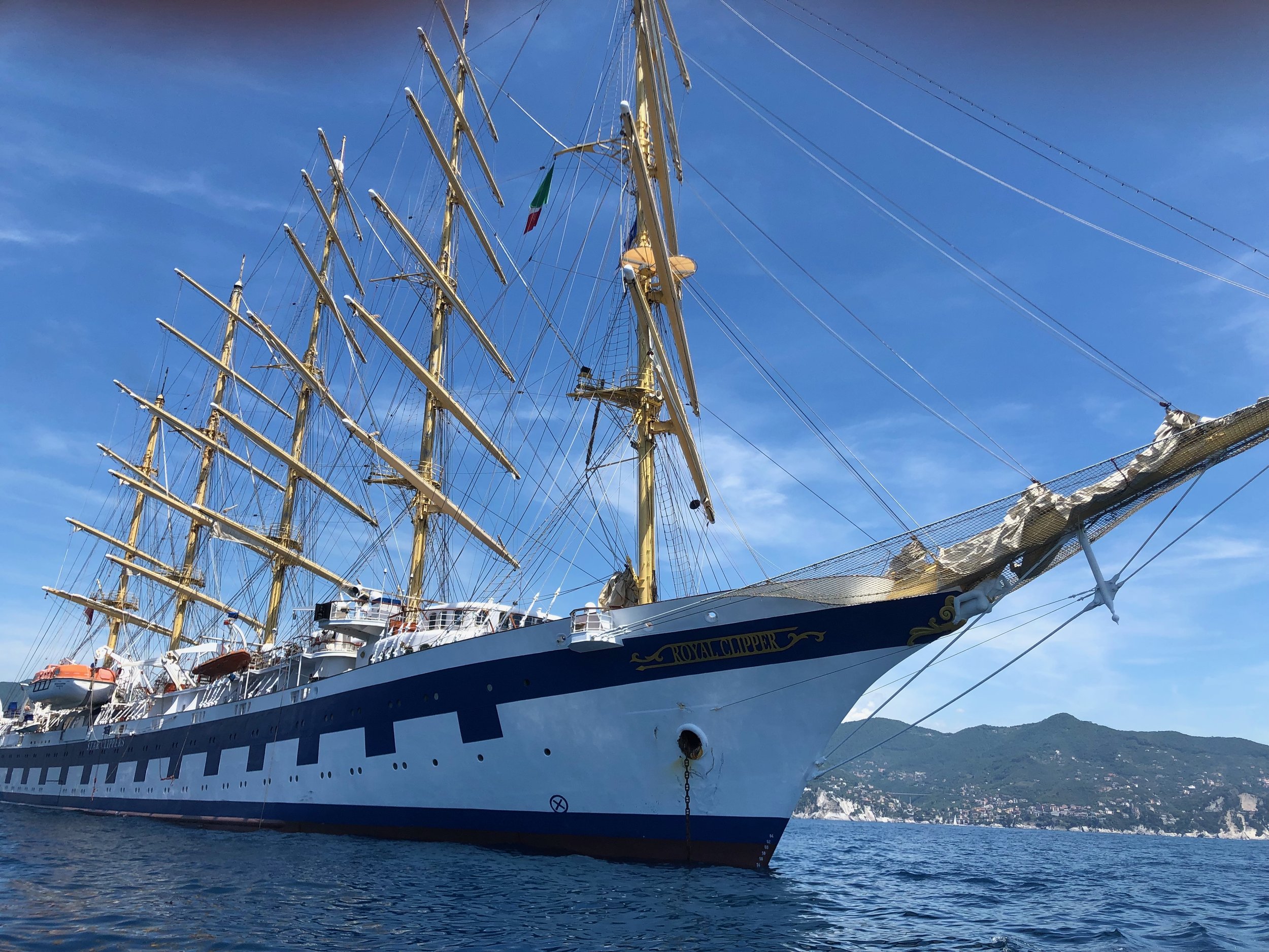 A large sailing ship named 'Royal Clipper' with multiple tall, yellow masts and rigging, anchored in a body of water with mountains in the background and a clear blue sky overhead.