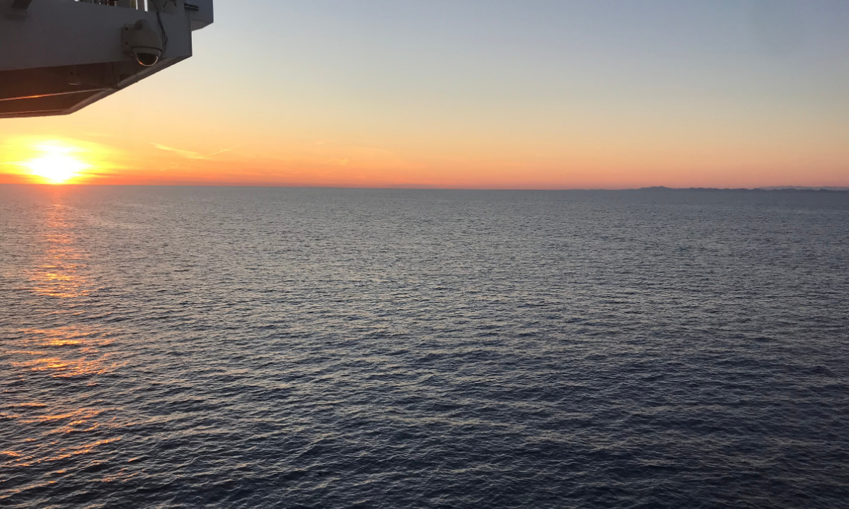 Sunset over the ocean as seen from a boat, with part of the boat's structure visible in the upper left corner.