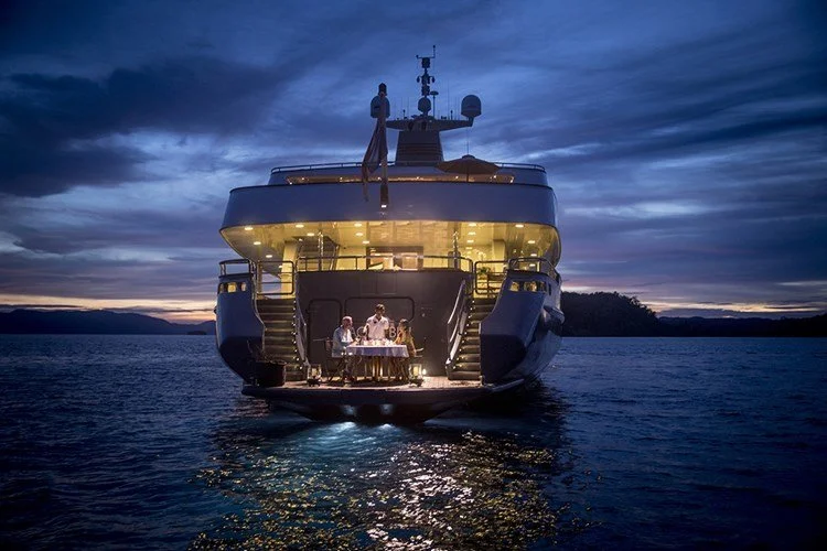 A yacht with people dining outside on a docked boat during twilight, with a dark water backdrop and cloudy sky.