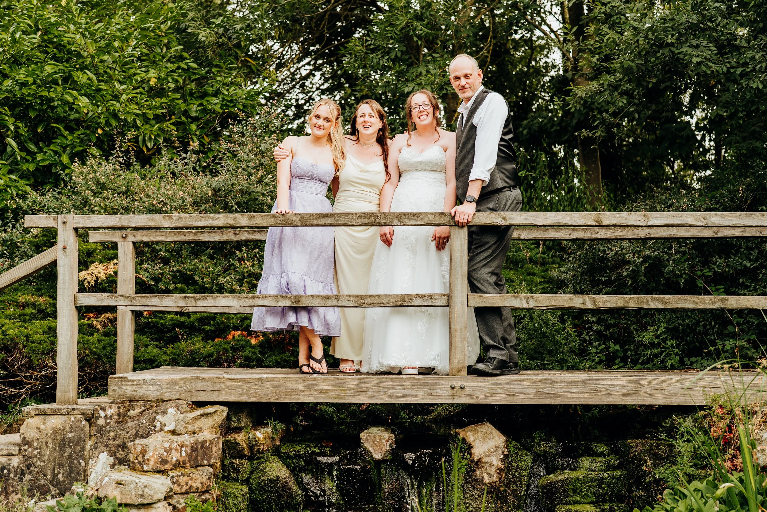 Four people standing on a wooden bridge surrounded by greenery. One person is wearing a wedding dress, three are in semi-formal attire, smiling and posing for the photo.