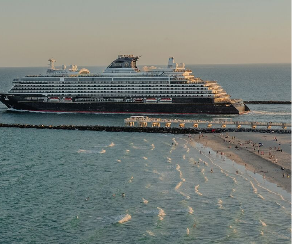 A large cruise ship passing by a sandy beach with people swimming and relaxing, with a pier and breakwater in the foreground.