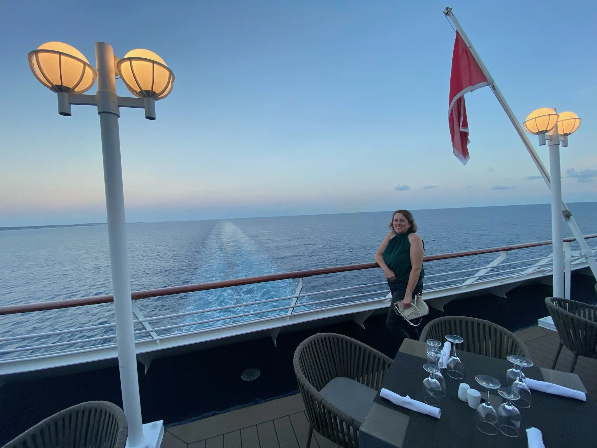 A woman smiling on the deck of a cruise ship, with table settings including glasses and napkins in the foreground and an ocean view in the background, during the evening with a clear sky.
