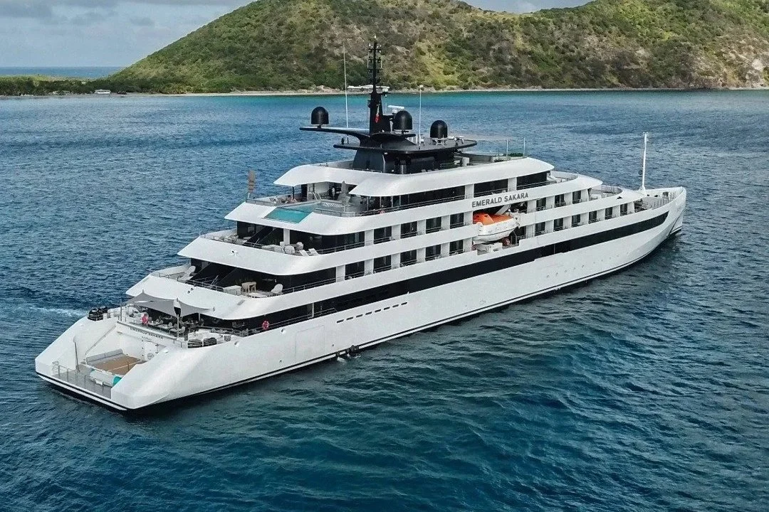 A large white yacht named Emerald Sakara sailing in the ocean with a green, hilly island in the background.