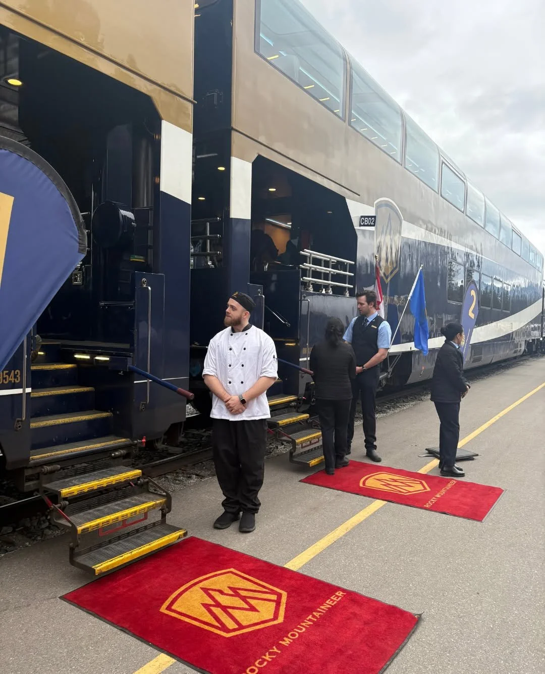 A large double-decker train decorated in blue and gold with the Rocky Mountaineer logo, lined up at a station with red Rocky Mountaineer branded rugs, staff members, and flags.