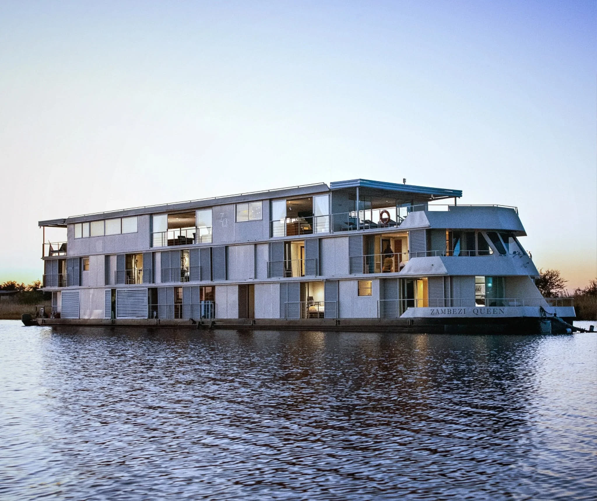 Modern houseboat named Zambezi Queen on a river at sunset, with large windows and outdoor balcony.