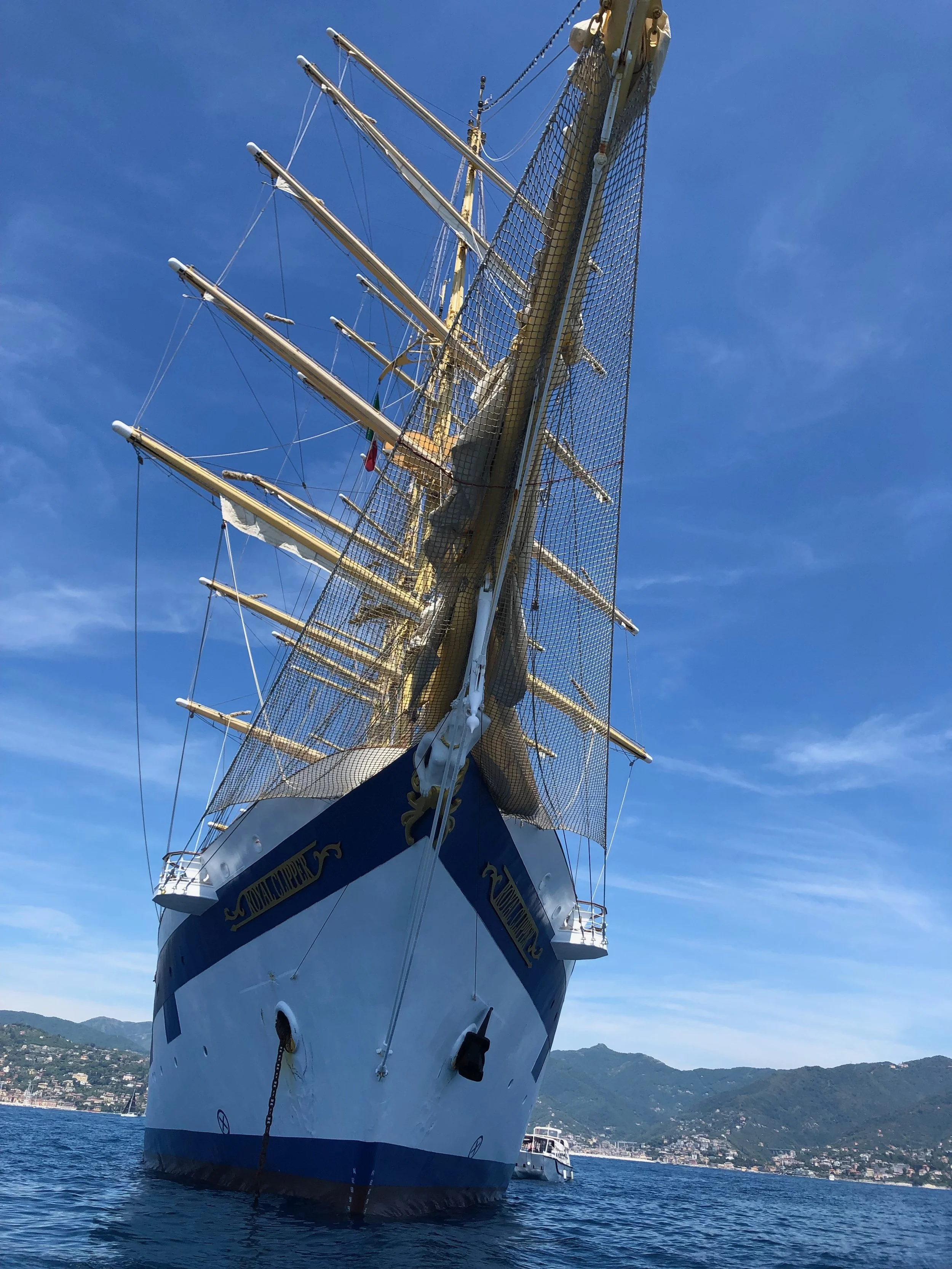 A large sailing ship with multiple masts and rigging docked in a harbor, with a city and mountains in the background and a clear blue sky above.