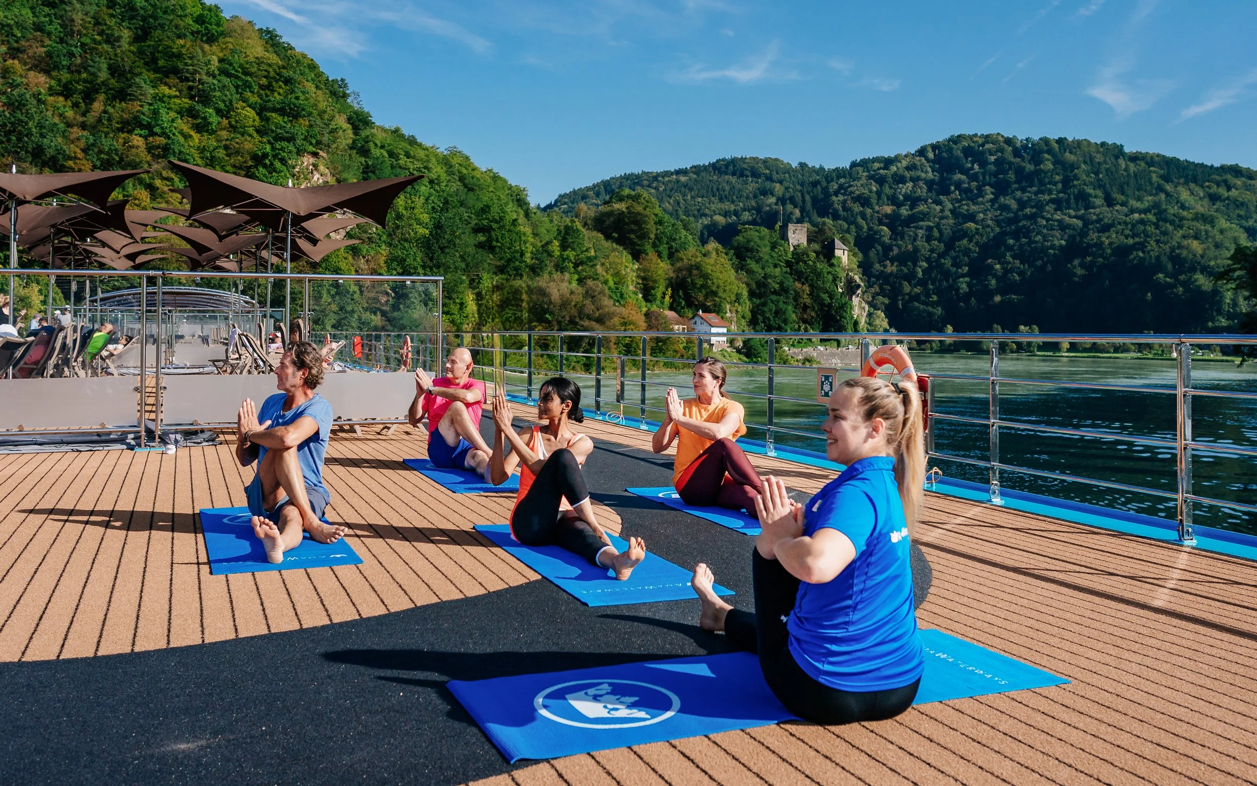 Group of people practicing yoga outdoors on a boat deck surrounded by water and green hills.