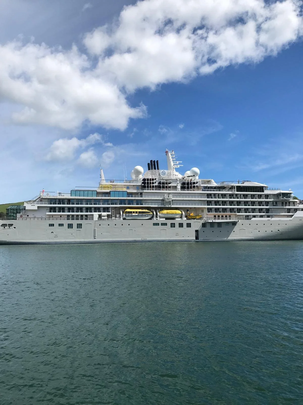 Large white cruise ship with yellow lifeboats docked on calm water under partly cloudy blue sky.