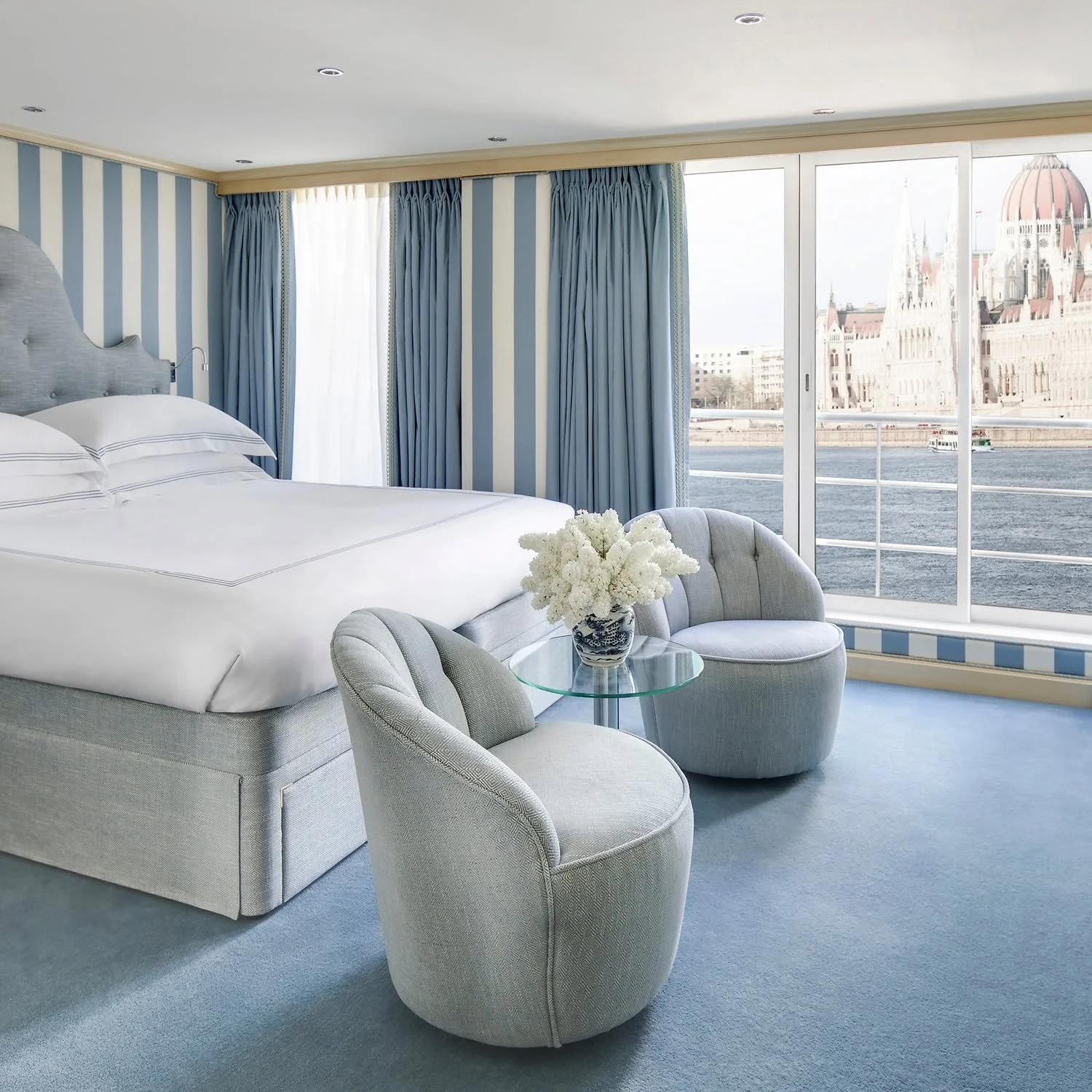 A hotel room with a large bed, light gray upholstered furniture, a glass-topped table with a flower vase, and floor-to-ceiling windows showing a cityscape and river view.