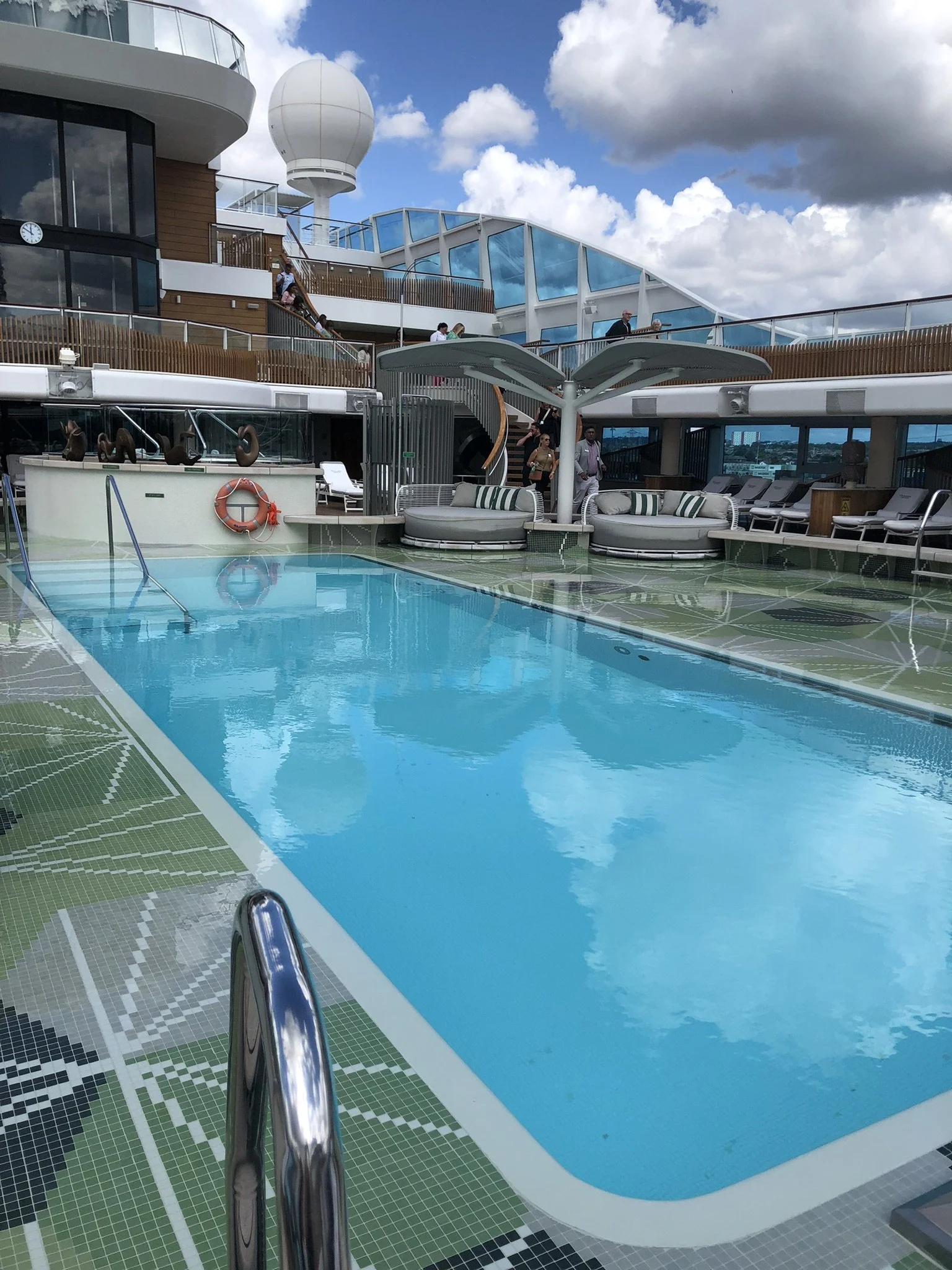 A view of a swimming pool on a cruise ship deck with a metal handrail leading into the pool, surrounded by lounge chairs and shaded seating area, with multiple levels of the ship's deck, people walking, a radar or weather sphere, and a cloudy sky in the background.