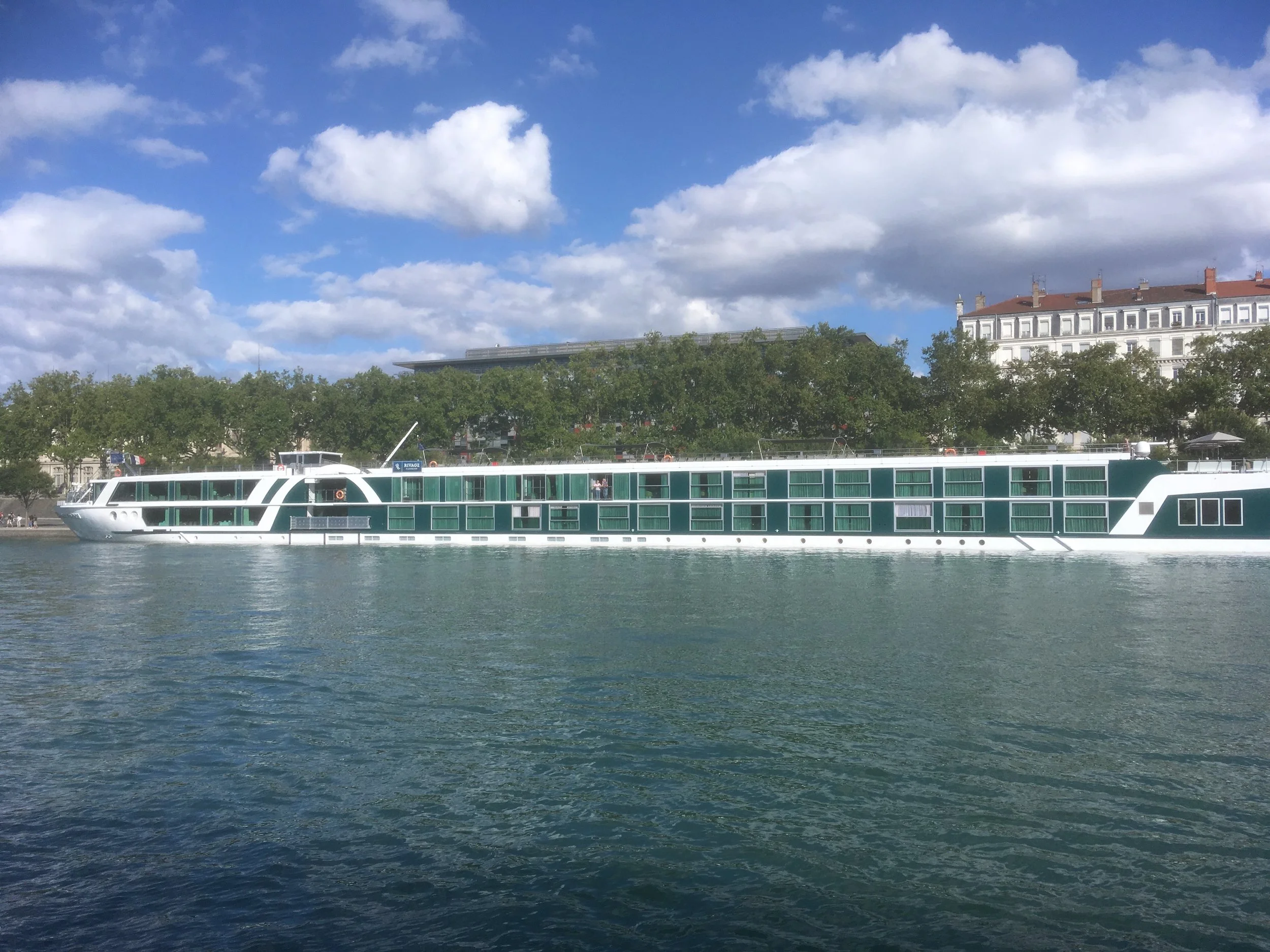A white and green boat sailing on a river with trees and white buildings in the background under a partly cloudy blue sky.