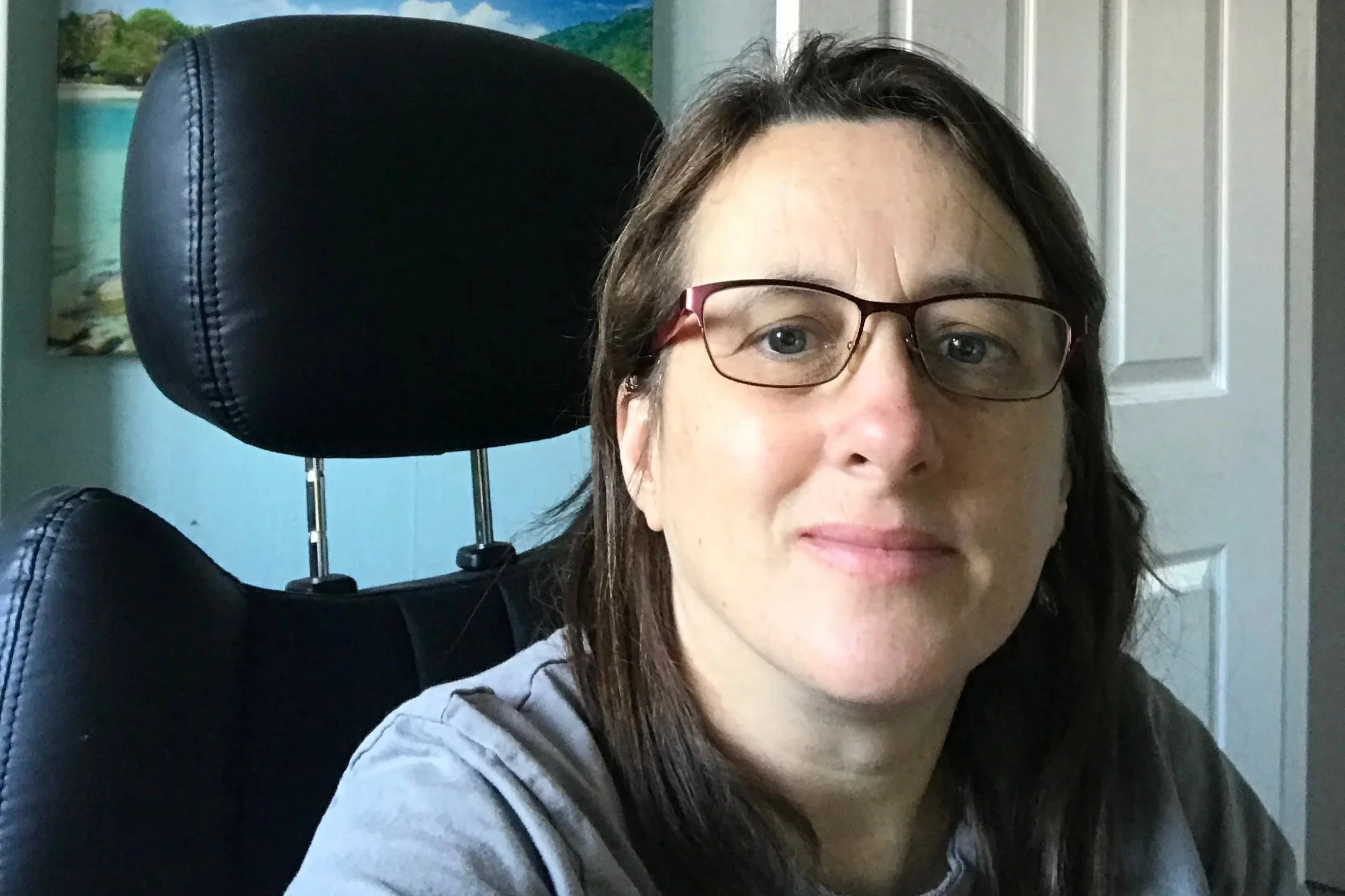 A woman with brown hair and glasses sitting in a wheelchair. She is smiling slightly. There is a landscape painting and a white door in the background.