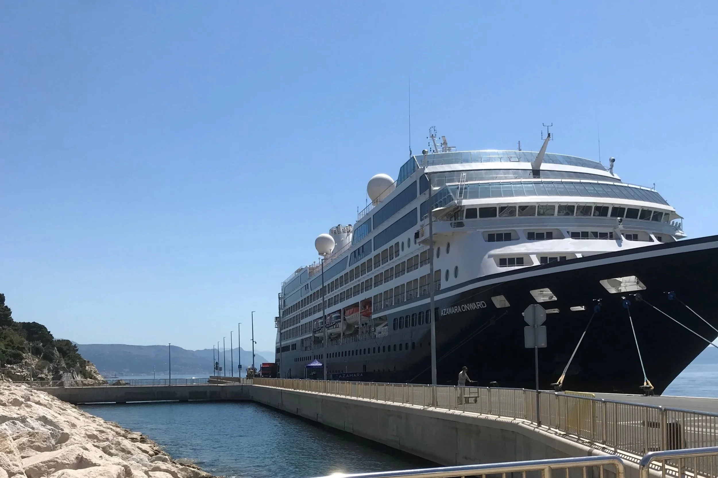 A large white cruise ship docked at a port with mountains in the background. The ship has multiple decks and is moored at a pier by a waterfront walkway, with a few people visible on the ground.