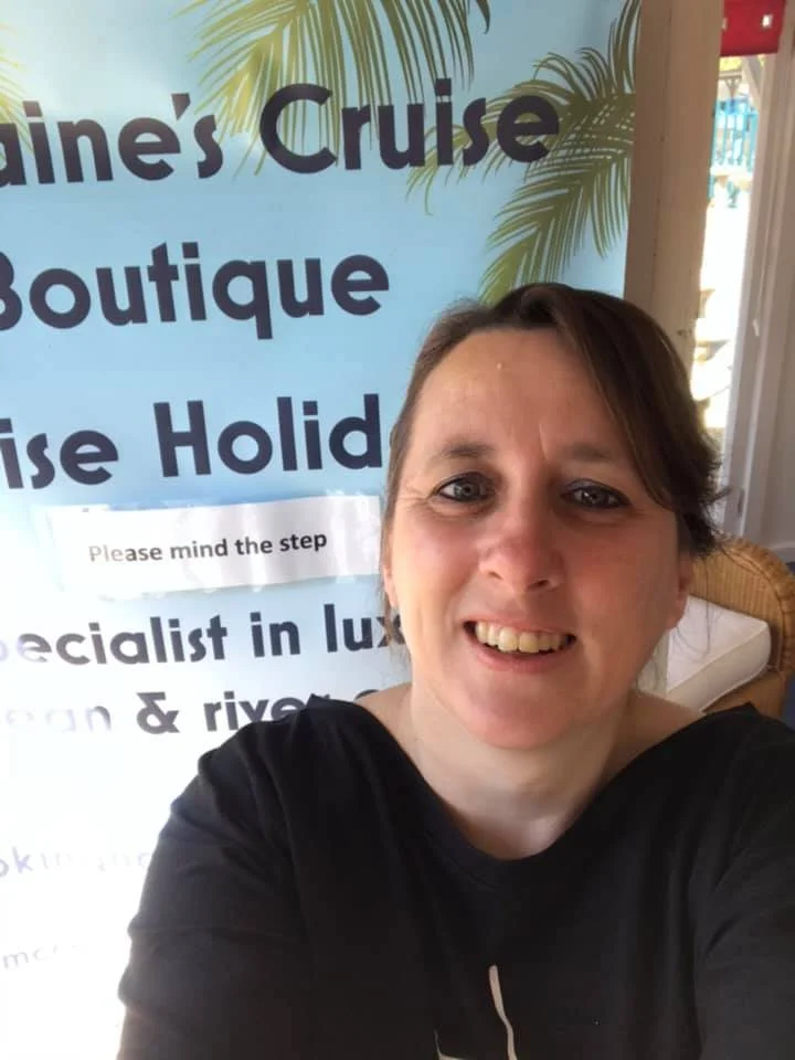 A smiling woman taking a selfie in front of a sign that reads "Lorraine's Cruise Boutique" with tropical palm tree graphics.