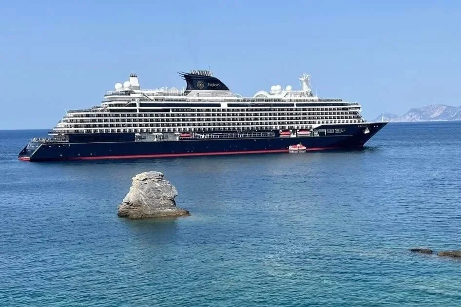 Large cruise ship with black and white exterior sailing in the ocean near rocky islands under clear blue sky.