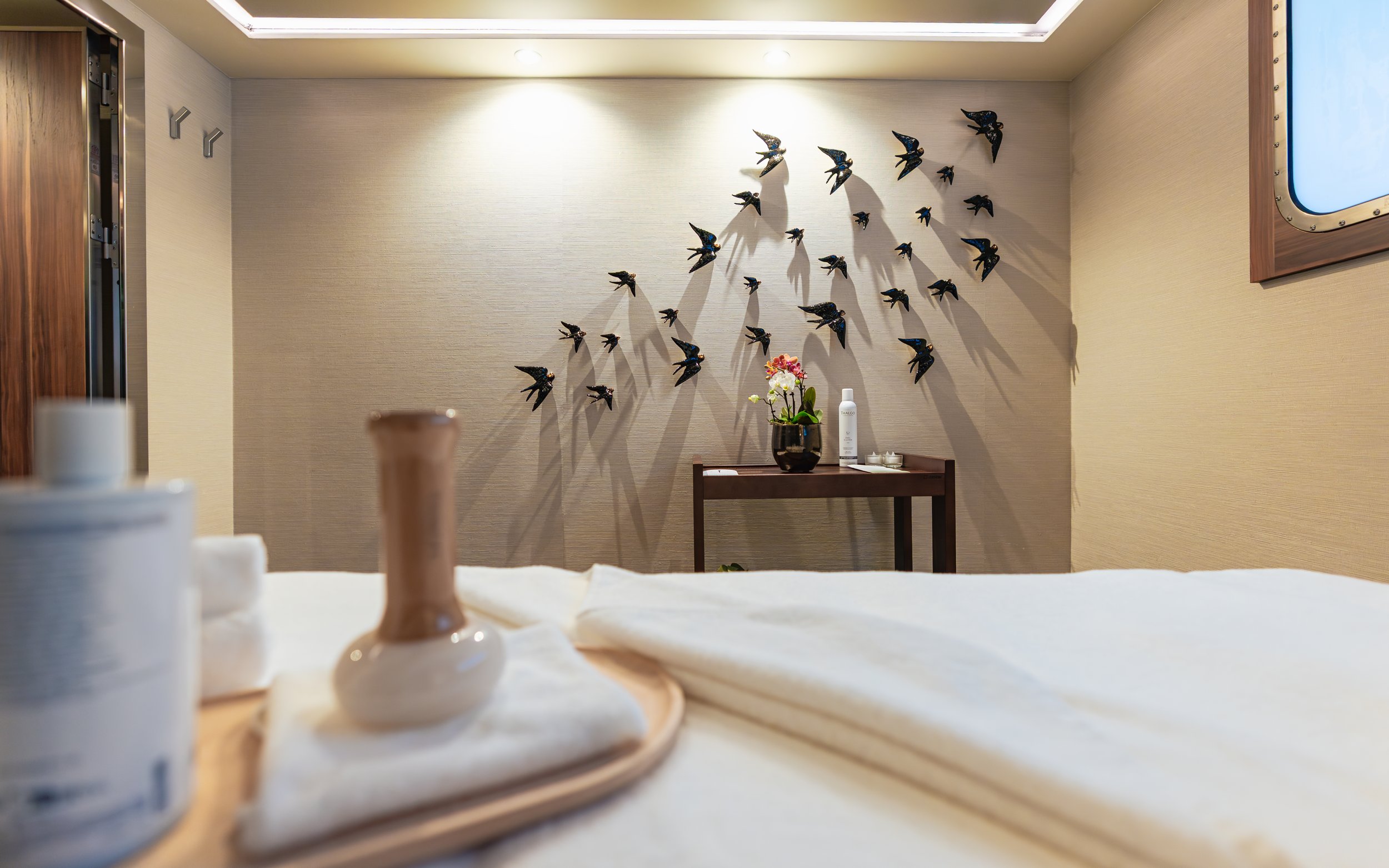 Spa room with a bed, a tray with a candle, a small vase, and a towel in the foreground; wall decor featuring multiple black bird sculptures shadows in the background; small table with a flower arrangement and toiletries; soft lighting.