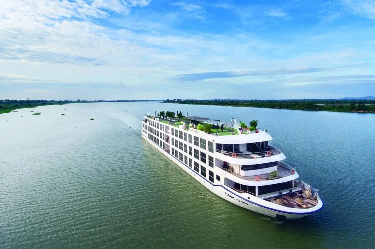 A large white cruise ship sailing on a wide river under a partly cloudy sky with green banks in the distance, featuring multiple decks and rooftop outdoor areas.