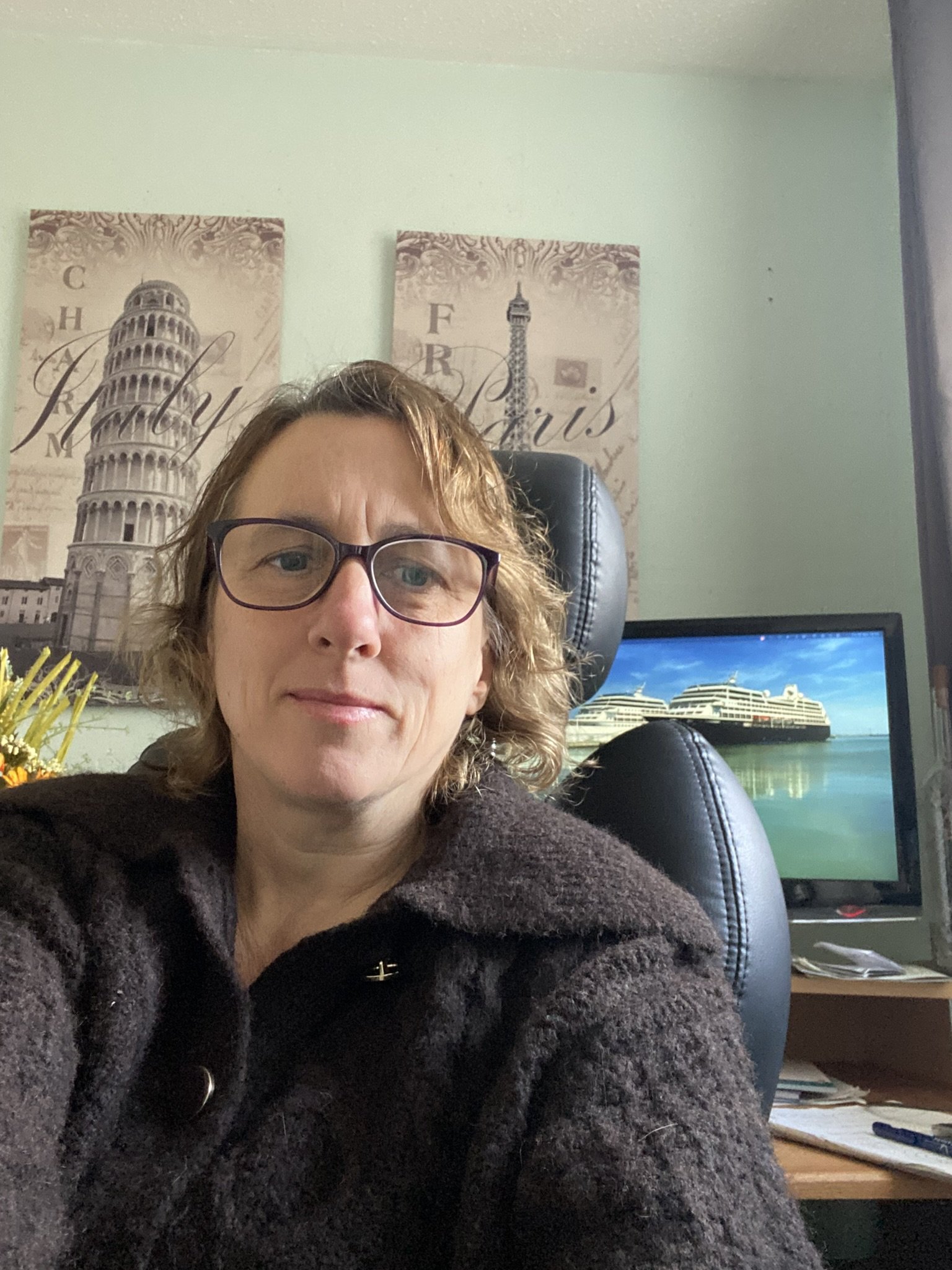 A woman with glasses taking a selfie in her office with paintings of the Leaning Tower of Pisa and the Eiffel Tower, and a computer screen showing a cruise ship, behind her.