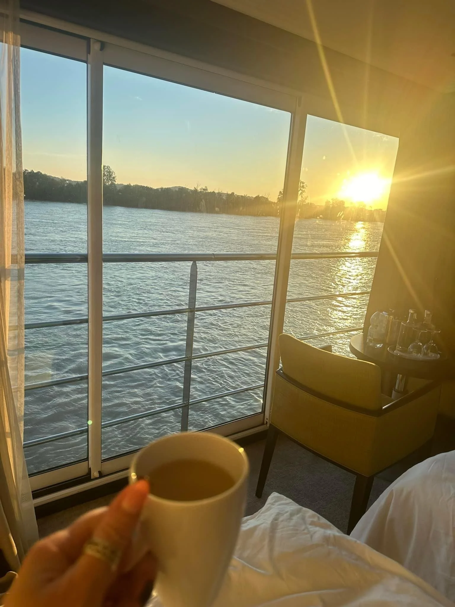 View from a hotel room with a balcony overlooking a river at sunset, person holding a coffee cup in the foreground, bed and chair visible inside the room.