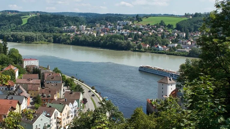 A scenic view of a river with a boat cruising along, surrounded by houses and greenery on both sides, with a town and rolling hills in the background.