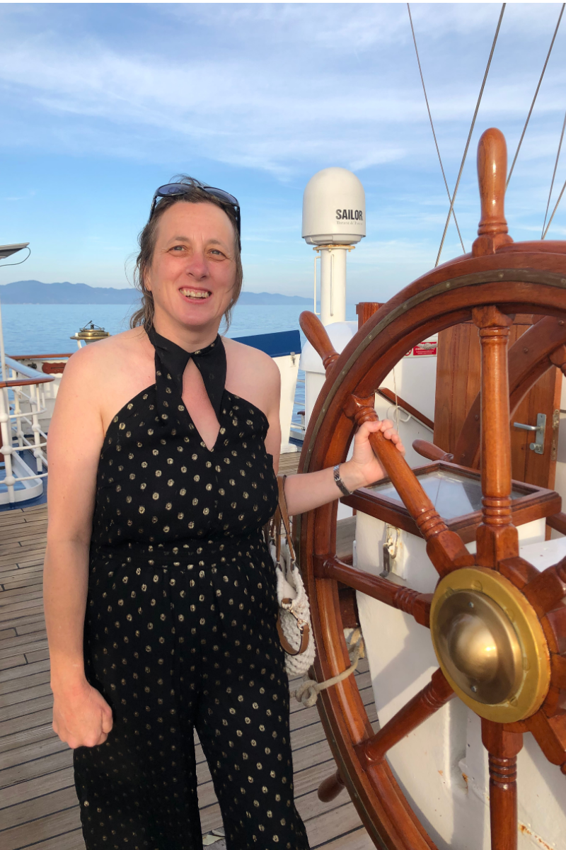A woman standing on a ship deck near the helm, smiling, with water and distant mountains in the background.