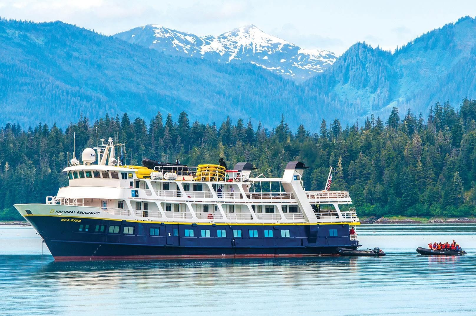 A large white and blue tour boat named Sea Bird on calm water with a forested shoreline and snow-capped mountains in the background. A smaller raft with people wearing orange life jackets is nearby.