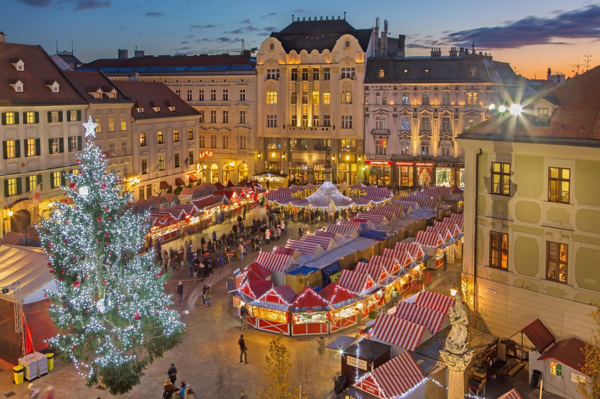 Christmas market in a city square during the evening, decorated with a large, lit Christmas tree and numerous red and white striped market stalls, with historic buildings in the background.