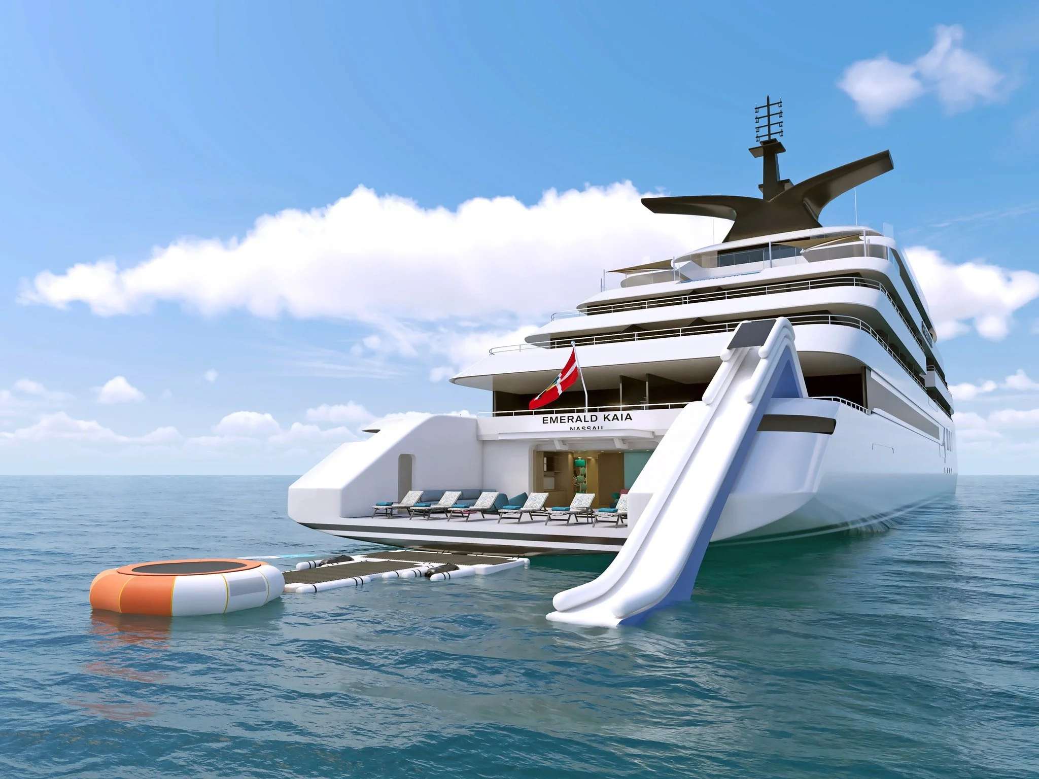 Luxury yacht named 'Emerald Kaia' in the ocean with a waterslide extending into the water, a floating trampoline, a flag, and an open sky with scattered clouds.