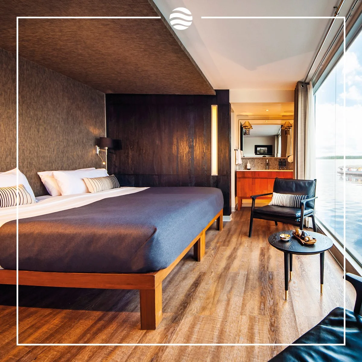 Modern hotel room with a large bed, black and white decor, wooden flooring, a black lounge chair with a pillow, a small round table with snacks, and large windows overlooking water.