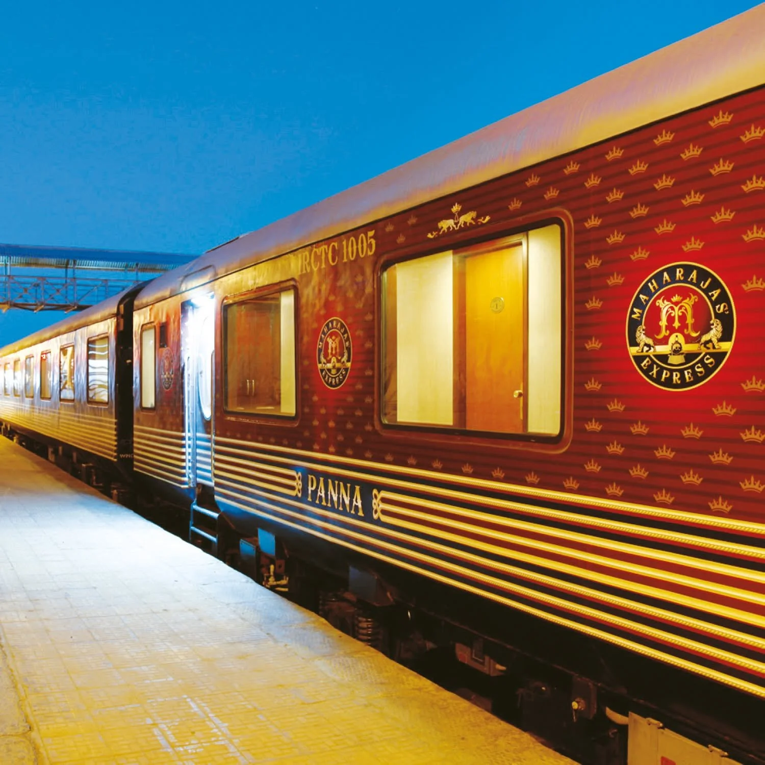 A luxury train painted in red with gold accents, featuring the Maharanjas Express logo and decorative crowns, parked at a station platform during evening.