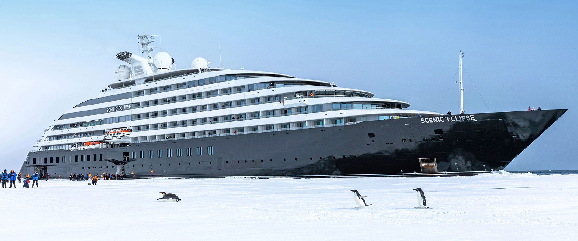 Large cruise ship named Scenic Eclipse on icy water, with penguins and people in winter clothing nearby.