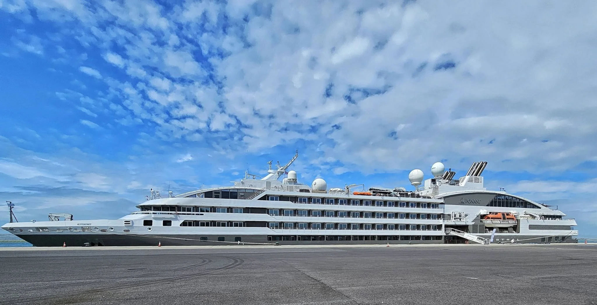 A large white luxury cruise ship docked at a port under a partly cloudy blue sky.