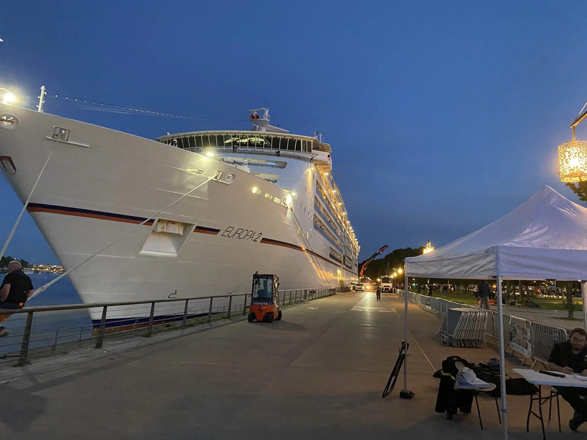Large white cruise ship docked at port during evening twilight with people sitting at tables, tents, and streetlights along the waterfront.