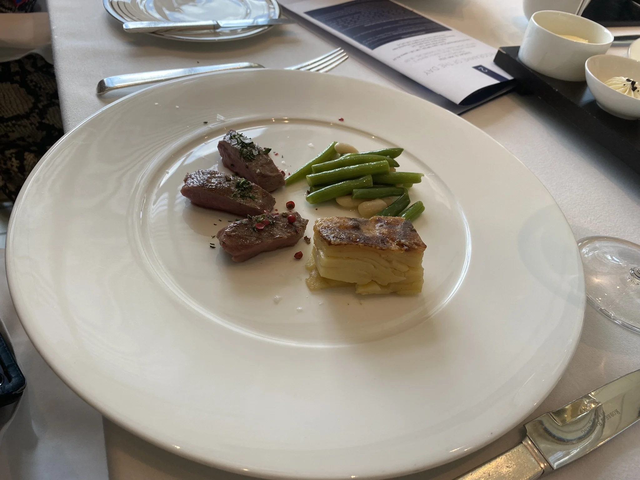 A white plate with slices of cooked steak garnished with herbs, green beans, white beans, a potato gratin, and a small pile of pink peppercorns, placed on a table with silverware, a menu, and small bowls in the background.